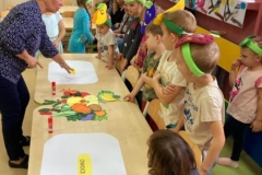 Grupa dzieci stoi za stolikami na których znajdują się dwie plansze z napisem: owoce i warzywa oraz powycinane, kolorowe owoce i warzywa. Dzieci przyklejają je na odpowiednią planszę.