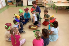 Dzieci siedzą z panią w kole na podłodze. Pani trzyma w ręku worek do którego dziewczynka wkłada rękę i zgaduję jaki owoc ma w ręce.