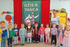 Grupa dzieci ubranych w kolorowe stroje stoją,  tworząc półkole.  W tle na zielonym płótnie widnieje napis Dzień  Tańca.8