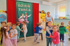 Dzieci stoją w parach twarzami do siebie trzymając się za ręce. Za nimi dekoracja z płótna oraz napis Dzień  Tańca.