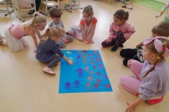 Grupa dzieci siedzi wokół niebieskiego kartonu odbijają dłonie pomalowane farbą.