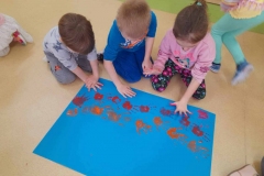 Na niebieskim kartonie troje dzieci odbija pomalowane dłonie farbą.