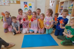 Wokół niebieskiego kartonu siedzą dzieci  kilkoro z nich ma pomalowane dłonie farbami.