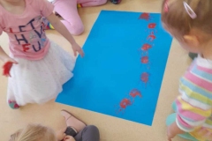 Grupa dzieci siedzi wokół niebieskiego kartonu odbijają dłonie pomalowane farbą.