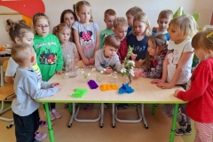 Dzieci zgromadzone wokół stołu, na stole stoją szklanki, kolorowa bibluła, w nich białe kwiatki.