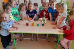 Dzieci zgromadzone wokół stołu, na stole stoją szklanki z zabarwioną wodą, w nich białe kwiatki.