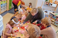 Przy stoliku siedzą 4 dziewczynki i 2 panie,  trzecia pani nachyla się nad stolikoiem. Dzieci smarują klejem drobne elementy i przyklejają na czerwonej kartce. 