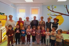 Grupa dzieci stoi w dwóch rzędach tworząc półkole. Każde z nich trzyma w rękach czerwone serduszko. Za nimi stoi siedem pań. W tle szara ściana.