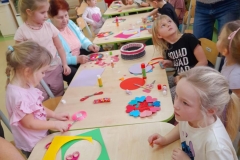 Dzieci siedzą przy stolikach z seniorami, wykonują walentynkę, na stołach leżą kolorowe materiały papiernicze.