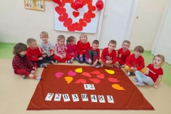 Dzieci ubrane na czerwono siedzą na podłodze, przed nimi na brązowym materiale poukładane połówki serc oraz różne cyferki.