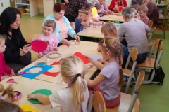 Dzieci siedzą przy stolikach z seniorami, wykonują walentynkę, na stołach leżą kolorowe materiały papiernicze.
