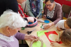 Dzieci siedzą przy stolikach z seniorami, wykonują walentynkę, na stołach leżą kolorowe materiały papiernicze.