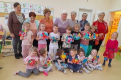 Kilkoro dzieci siedzi na podłodze, za nimi stoją inne dzieci, każde z nich trzyma w ręku Walentynkę. W tyle za dziećmi stoją Seniorzy.