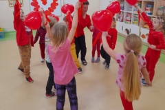 Dzieci ubrane na czerwono tańczą do muzyki, każde z nich trzyma w ręku czerwony balon.