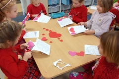 Sześć dziewczynek siedzi przy  stoliku i wycina serduszka.
