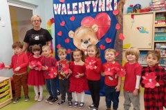 Dzieci ubrane w czerwone stroje stoją  w  szeregu. Każde trzyma w rączce czerwony kwiatek w kształcie serduszka. Za nimi  napis Walentynki na ciemnoniebieskim tle.