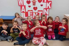 Grupa dzieci ubrana w czerwone stroje siedzi na podłodze.  Za nimi  na białym tle napis Walentynki.