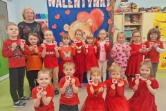 Pięcioro dzieci dzieci siedzi na podłodze. Dziesięcioro stoi tworząc  półkole.  Dzieci  ubrane w czerwone stroje,  każde z nich trzyma w rączce czerwonego lizaka w kształcie serce. Z tyłu  napis Walentynki.