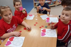 Grupa dzieci ubrana w czerwone stroje siedzi wokół stołu.  Na stoliku leżą czerwone serduszka. W tle okna.194482332709_n