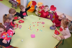 Grupa dzieci siedzi przy sześciokątnym stoliku. Każde dziecko nakleja papierowe elementy na kartonowy kształt czerwonego serca. Niektóre z nich trzymają w rękach gotowe ozdobione serca- walentynki. Za dziećmi widać przedszkolne mebelki i zabawki
