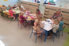 Dzieci siedzą przy 3 stolikach: niebieskim, fioletowym i zielonym. Przy pierwszym pani w kolorowej bluzce pomaga siedzącemu obok chłopcu wybrać kolor kredki. Przy stoliku zielonym pani w białym żakiecie pokazuje chłopcu w niebieskiej bluzie, co pomalować kolorem czerwonym.