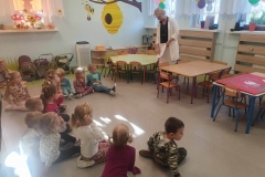 Dzieci siedzą na podłodze. Przy zielonym stoliku stoi pani w biało- czarnym ubraniu. W rękach trzyma kartkę papieru. W tle zawieszone na firankach kolorowe balony, na parapetach ustawione kwiaty.