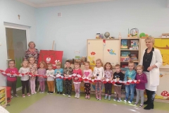 Grupa dzieci stoi w półkolu. W rękach trzymają biało- czerwony łańcuch. Obok nich stoją 2 panie. Za nimi dekoracja na Święto Niepodległości: biały orzeł w złotej koronie na tle drzewa. Obok napis: 11 LISTOPADA. Dookoła łańcuch z biało- czerwonych harmonijek.