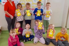 Kilkoro dzieci stoi wraz z wychowawczynią na tle tablicy, parę dzieci siedzi na podłodze, każde z nich trzyma w ręku żółte Misie.