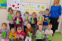 Kilkoro dzieci stoi na tle tablicy z panią Jadzią, parę dziedzi na podłodze, każde z nich trzyma w ręku kolorowa choinkę.