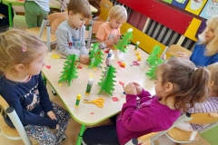 Czwórka dzieci siedzi przy stoliku, ozdabia zielone choinki, pomaga im pani z GOK.