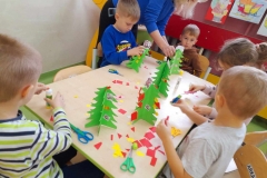 Piecioro dzieci siedzi przy stoliku, ozdabia zielone choinki, pomaga im pani z GOK.