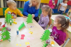 Dwie dziewczynki I chłopiec siedzą przy stoliku, ozdabiają zielone choinki, pomaga im pani z GOK.