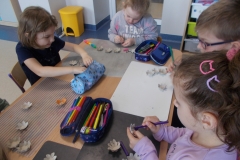 Przy stoliku przedszkolnym siedzą trzy dziewczynki i jeden chłopiec. Dzieci kolorują pisakami wycięte z kartonu kwiaty. Leżą one na plastikowych szarych podkładkach. Na stole znajdują się trzy kolorowe piórniki. W tle żółty kosz na śmieci i szafki.