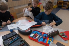 Przy stoliku przedszkolnym siedzi trzech chłopców ubranych w ciemne bluzy. Jeden z nich zwija wzorzystą serwetkę w harmonijkę, drugi trzyma ją w ręce. Trzeci koloruje szare, kartonowe kwiatki. Na stole leżą piórniki z przyborami pisarskimi.