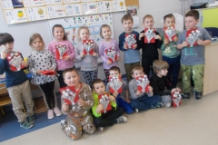 Grupa dzieci przedszkolnych demonstruje swoje prace plastyczne - bukiety kwiatów na czerwonych kartkach. W tyle stoi dziewięć dzieci, z przodu siedzi pięcioro. Ubrane są w kolorowe stroje. Stoją one na tle ściany, na której znajdują się obrazki z literami oraz obrazki tematyczne do widocznego napisu: ,, zdrowie’’.