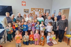 Grupa dzieci stoi w półkolu. Przed nimi na beżowej podłodze klęczy 7 dzieci. W rękach trzymają kolorowe koszyczki z kartonu, a w nich kwiaty z krepiny. Za dziećmi stoją dorośli. Na ścianie wisi godło, krzyż, telewizor, tablica korkowa i 2 obrazy.