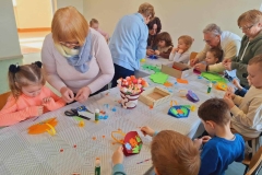  Dzieci siedzą przy stolikach. Przed nimi leżą koszyczki z kolorowego kartonu. Naklejają na nich kwiaty, trawę, listki. Nad dziećmi pochyla się 5 pań. Pomagają wycinać ozdobne elementy.