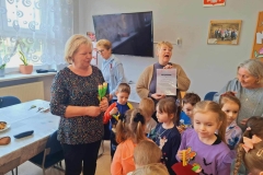  Pani w beżowym swetrze trzyma w rękach "Podziękowanie" od przedszkolaków. Przed nią stoją dzieci. Obok nich 3 panie. Jedna w ciemnej bluzce ma 2 kwiatki z krepiny. W tle okno z firanką.