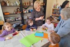 Czworo dzieci siedzi przy stole, na którym leżą kolorowe papiery, klej, nożyczki, koszyczki z kartonu. Przy nich stoją 3 panie. W tle regały, na których ustawione są gotowe prace, m.in.kwiaty, ozdoby wielkanocne, jeż z szyszek oraz pudełka z przyborami plastycznymi.