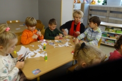 Przy stoliku przedszkolnym siedzi czworo dzieci. Obok nich przykucnęła kobieta     w ciemnym stroju i czerwonym szaliku. Pomaga dzieciom w tworzonych pracach plastycznych. Na stoliku obok ścinków papieru leżą kleje i nożyczki. Z prawej strony widać częściowo głowy dwóch dziewczynek. W tle półka z układankami.