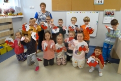 W sali przedszkolnej na tle ściany i tablic widać stojące i klęczące dzieci wraz ze stojącą za nimi wychowawczynią. Dzieci ubrane są w kolorowe stroje.  Trzymają           w rękach wykonane przez siebie prace plastyczne: biało czerwone rozetki z orłem na środku oraz biało czerwone sercowe kwiaty też z orłem w środku. Z boku widoczne żółte pojemniki z klockami.