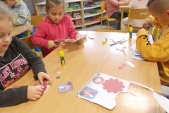 Przy stoliku siedzą dwie dziewczynki i chłopiec. Dziewczynka w różowej bluzie         i chłopiec w żółtej bluzie wycinają elementy z kartki, dziewczynka w ciemnej bluzie     z nadrukiem nakleja wycięte elementy. W tle częściowo widać jeszcze dwa stoliki        z pracującymi dziećmi oraz szafkę z układankami.