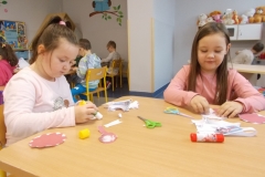 Przy stoliku przedszkolnym siedzą dwie dziewczynki. Jedna jest ubrana w białą, druga w różową bluzkę. Przyklejają wycięte z papieru elementy, tworząc biało czerwone emblematy. Na stoliku leża nożyczki, klej oraz ścinki z kartek. W tle widać inne dzieci siedzące przy stolikach. Nad nimi na ścianie namalowana jest sowa na gałęzi. Z prawej strony widoczny kącik kuchenny i bawiące się w nim dziecko.