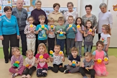 Grupa  dzieci stoi w półkolu za nimi stoją seniorzy. W pierwszym rzędzie 7 dzieci siedzi po turecku.