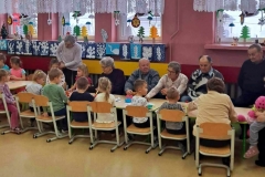 Dzieci siedzą przy stolikach, kleją laurki, Seniorzy im pomagają.