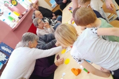 Dzieci siedzą przy stolikach, kleją laurki, Seniorzy im pomagają.