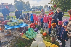 Grupa dzieci z 6 paniami stoi przed straganem z warzywami. Leżą na nim: kalafiory, cebula, koperek, pory, czosnek. Przy straganie kapusta biała, czerwona,   włoska, dynia oraz worki z kapustą szatkowaną i ziemniakami. Za straganem pan w czerwonej czapce trzyma przy uchu telefon. Nieco dalej stoi 2 panów. W tle budynek banku i drzewa.