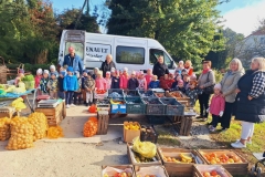 Grupa dzieci w kolorowych kurtkach i czapkach na głowach stoi w półkolu na tle białego samochodu dostawczego. Z lewej strony pan w dżinsowej kurtce i czerwonej czapce. Nieco dalej 2 panie w czarnych kurtkach. Z prawej strony pani, która trzyma na rękach dziewczynkę. Przy niej pani w szarej kurtce trzyma za rękę chłopczyka. Obok pani, przed którą stoi dziewczynka w różowej kurtce. Na końcu pani w koszuli w biało- czarną kratę i czarnej kamizelce. Przed dziećmi na straganie skrzynki ze śliwkami, winogronem, mandarynkami i bananami. Na ziemi pudełko z cytrynami i koszyk ze słoikami miodu. Nieco dalej skrzynki z jabłkami i gruszkami. Z lewej strony worki z ziemniakami.