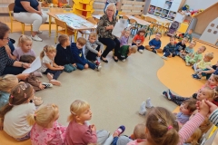 Grupa dzieci siedzi w kole na szaro- żółtej podłodze. Między nimi 2 panie w okularach: jedna siedzi na podłodze, druga na krzesełku. Obydwie trzymają w rękach białe kartki. Za nimi stolik,  przy którym siedzi pani w białych spodniach. Na stoliku leżą kartki przymocowane do patyczków z namalowanymi warzywami.