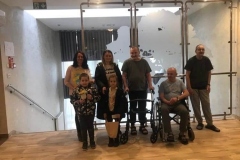 Zdjęcie przedstawia pięć osób dorosłych oraz dwoje dzieci na tle szklanej ściany. Mężczyzna siedzi na wózku. Pozostałe osoby stoją.